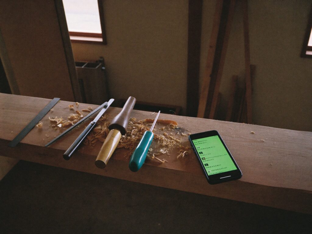 木くずの残る作業台に並ぶ大工道具と、チャット画面を開いたスマートフォン